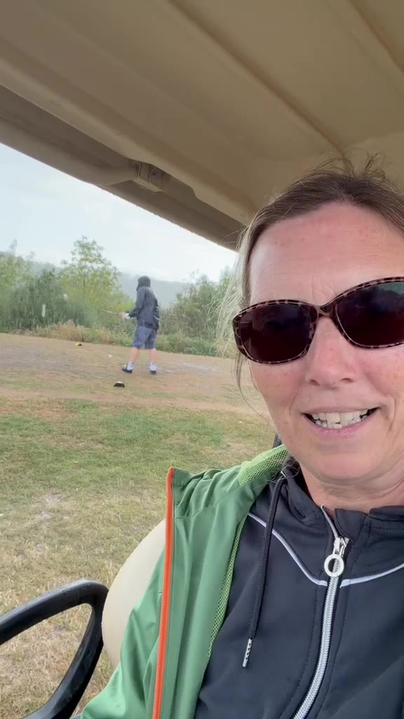 Lisa in sunglasses smiling in a golf buggy while Brian stands on the tee behind her