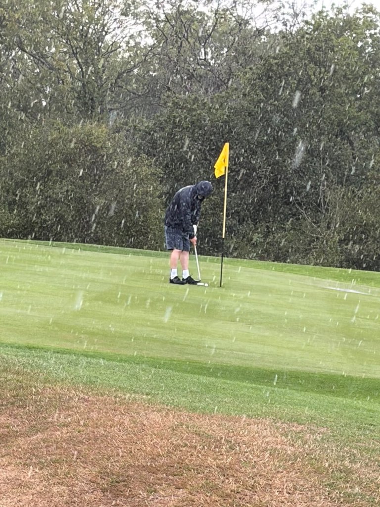 Brian putting in heavy rain at Brynhill, wearing shorts and a hooded jacket, beside a yellow flag
